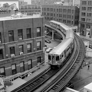 May include: A black and white photograph of a train traveling on a curved track in an urban setting. The train is a subway car with multiple windows and a door on the side. The train is traveling on a track with a concrete platform and a metal fence. The train is traveling through a city with buildings in the background.