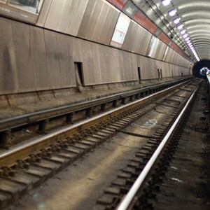 Puede incluir: Vista de un largo túnel de tren oscuro con dos vías. El túnel está revestido de azulejos blancos y tiene una serie de luces en el techo.