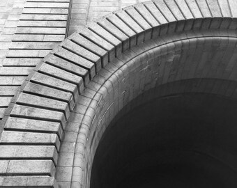 May include: Low-angle, black and white photograph of a stone archway and building facade. The archway is constructed of large, curved stones, and the building features rectangular windows and a textured surface. The image emphasizes architectural details.