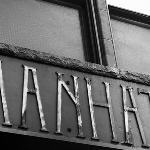 May include: A black and white photo of a building with the word "MANHATTAN" written in large, silver letters on the side.