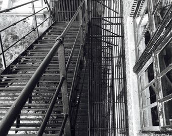 Könnte beinhalten: Schwarz-Weiß-Bild einer äußeren Metall-Feuerleiter. Die Feuerleiter hat Treppen, Geländer und einen vertikalen Leiterabschnitt. Das Backsteingebäude hat Fenster.