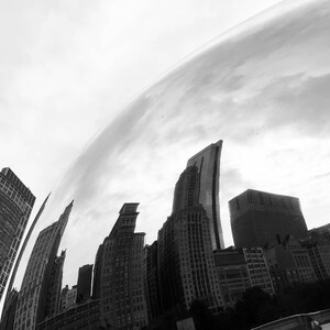 Könnte beinhalten: Ein Schwarzweißfoto einer großen, reflektierenden, silbernen Kugel, die die Skyline der Stadt widerspiegelt. Die Kugel befindet sich im Vordergrund und die Gebäude im Hintergrund. Die Gebäude sind hoch und schlank, mit vielen Fenstern. Der Himmel ist bewölkt.