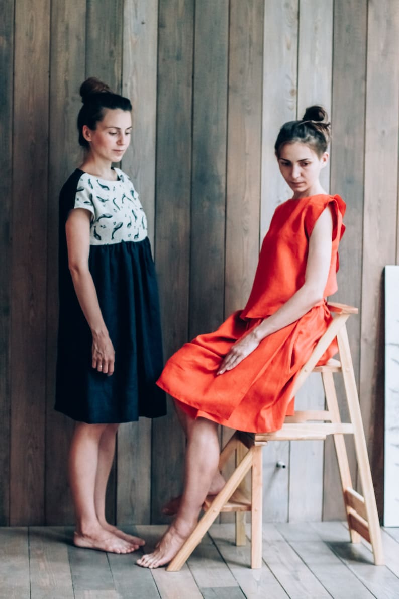 Peut inclure: Deux femmes portant des robes, l'une en robe bleu marine avec un motif blanc et noir, l'autre en robe rouge. La femme en robe rouge est assise sur un tabouret en bois.