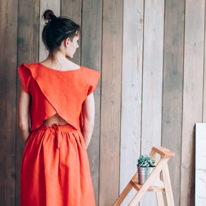 Peut inclure: Une femme portant une robe en lin rouge avec un dos d&eacute;gag&eacute;. La robe a un col carr&eacute; et un corsage ajust&eacute;. La jupe est ample et descend jusqu'aux genoux. La femme est debout devant un mur en bois.