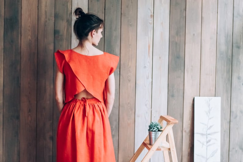 Peut inclure: Une femme portant une robe en lin rouge avec un dos d&eacute;gag&eacute;. La robe a un col carr&eacute; et un corsage ajust&eacute;. La jupe est ample et descend jusqu'aux genoux. La femme est debout devant un mur en bois.