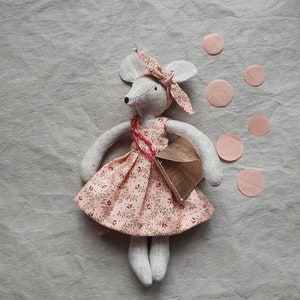 Puede incluir: Una muñeca de ratón gris hecha a mano que lleva un vestido rosa floral y un bolso marrón. La muñeca está sentada sobre un fondo de tela gris claro con círculos de papel rosa esparcidos a su alrededor.