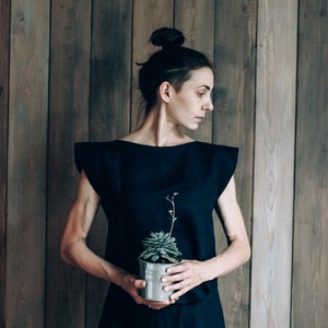 May include: A woman in a black dress holds a small potted succulent plant in front of a wood panel wall.