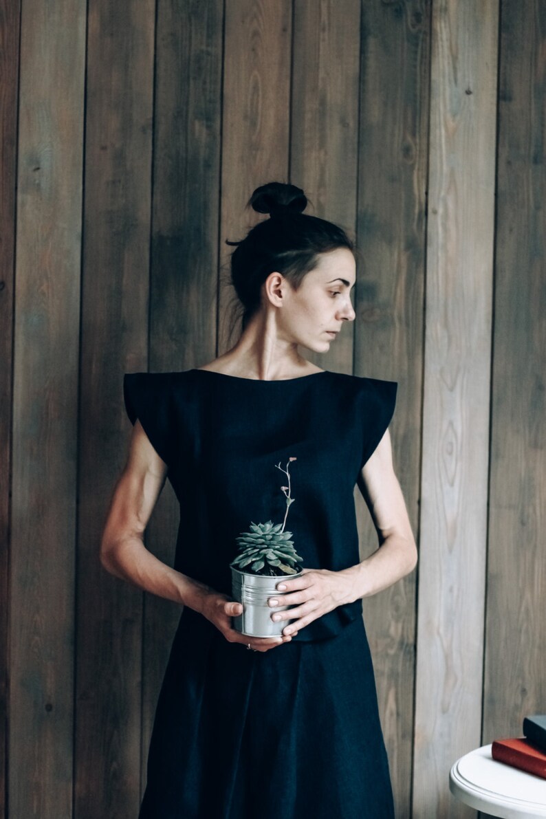 May include: A woman in a black dress holds a small potted succulent plant in front of a wood panel wall.