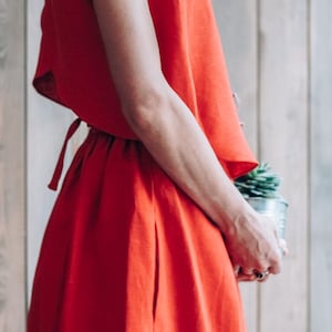 Peut inclure: Une femme portant une robe en lin rouge avec un lien &agrave; la taille. Elle tient une petite plante en pot.