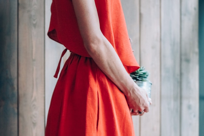 Peut inclure: Une femme portant une robe en lin rouge avec un lien &agrave; la taille. Elle tient une petite plante en pot.