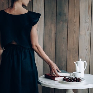 May include: A woman wearing a black dress stands in front of a wood panel wall. A white table with a teapot, a cup, a book, and a bowl of cherries is in front of her.