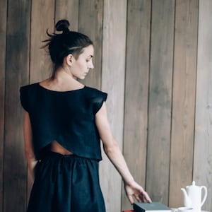 May include: A woman wearing a black linen dress with a low back, standing in front of a wood panel wall. She is reaching for a stack of books on a white table with a bowl of cherries.