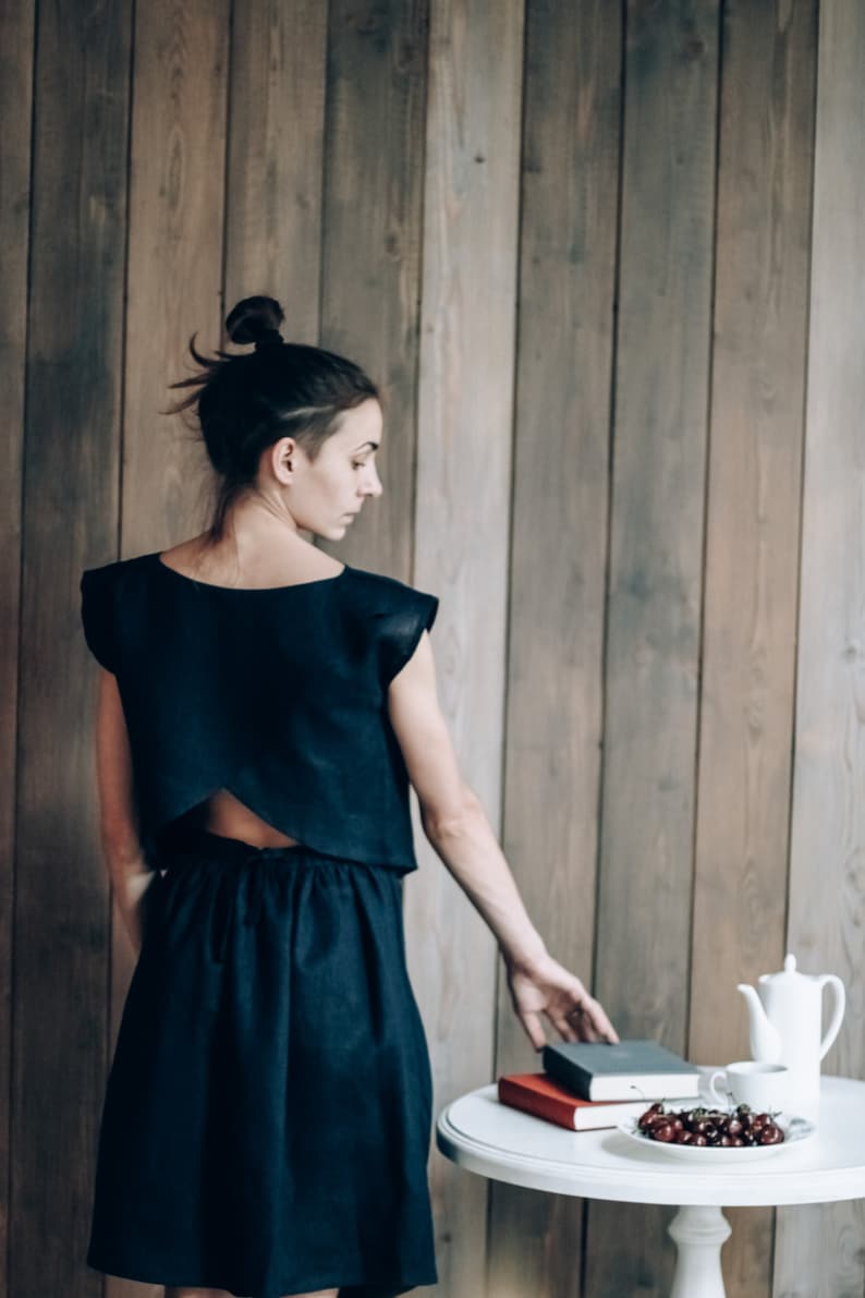 May include: A woman wearing a black linen dress with a low back, standing in front of a wood panel wall. She is reaching for a stack of books on a white table with a bowl of cherries.