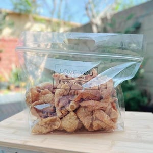 May include: A clear plastic bag filled with golden-brown, crispy fried snacks. The bag has a handle and a printed label that reads "LOVE YOU With KINDNESS Enjoy your life". The bag is sitting on a light-colored wooden surface.