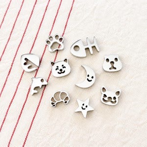 May include: A collection of small, silver-colored charms in various shapes, including a paw print, fish bone, cat, dolphin, croissant, moon, star, and two different dog faces. The charms are arranged on a white and red striped fabric.