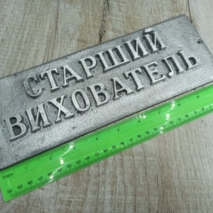 Puede incluir: Un letrero de metal plateado con el texto "Старший Вихователь" en letras negras. El letrero mide aproximadamente 25 cm de largo y 7,5 cm de ancho.