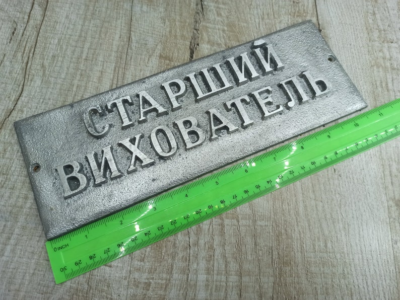 Puede incluir: Un letrero de metal plateado con el texto "Старший Вихователь" en letras negras. El letrero mide aproximadamente 25 cm de largo y 7,5 cm de ancho.
