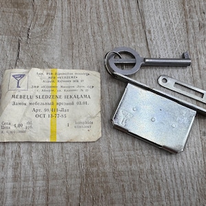 May include: An antique metal furniture lock set with a key, strike plate, and a paper label. The key is silver-colored, and the lock has a rectangular shape. The label has text in a foreign language.