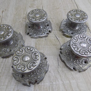 May include: Five silver door knobs with ornate designs. Each knob has a round, raised center with a floral pattern and is attached to a square base with intricate scrollwork.