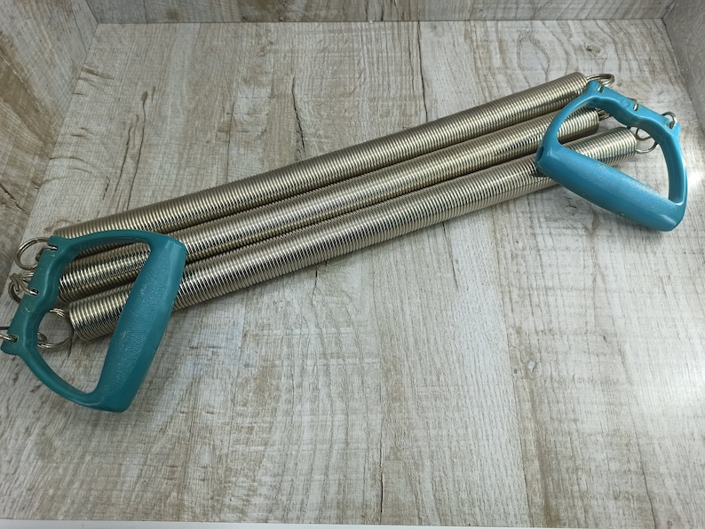 May include: A silver exercise expander with two teal plastic handles. The expander is made of multiple coiled springs.