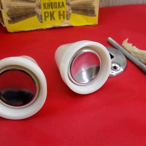 Può includere: Due pomelli per porte in ceramica bianca con un centro argentato. I pomelli sono su una superficie rossa.