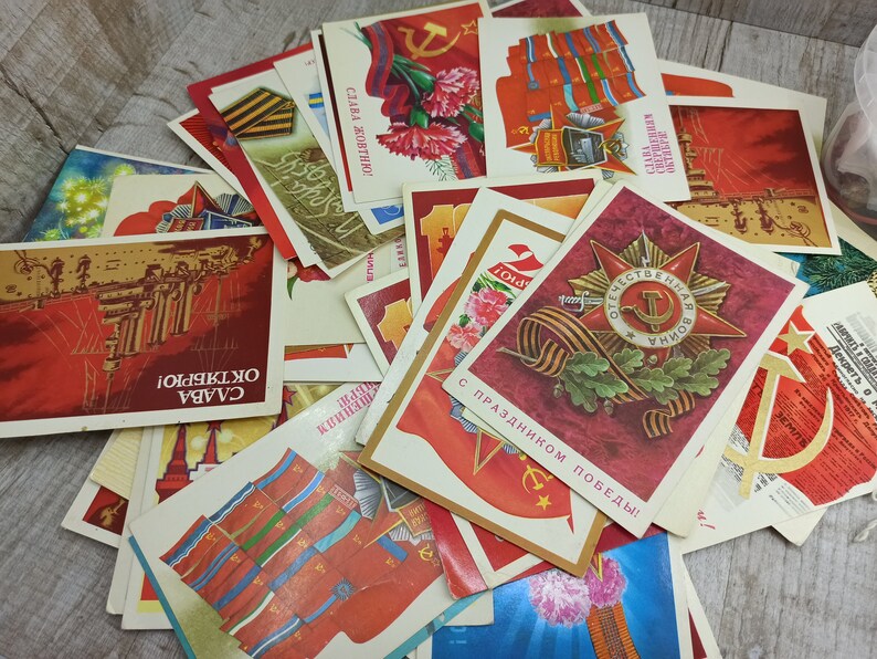 May include: A collection of vintage postcards with Soviet-era imagery. The cards feature red and gold designs, including military medals, floral arrangements, and the hammer and sickle symbol. Some cards have Cyrillic text.
