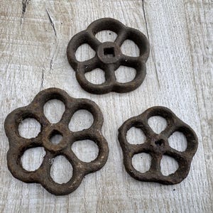 May include: Three antique cast iron valve handles in different sizes. Each handle features a flower-like design with open spaces. The metal has a dark, rusty brown colour, indicating age and outdoor use. They are placed on a weathered, light-coloured wooden surface.