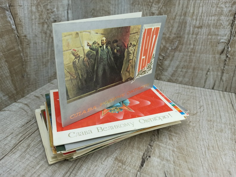May include: A stack of vintage postcards featuring Soviet-era imagery. The top card has a silver border and a painting of a man addressing a crowd. Other cards display red stars and Cyrillic text.