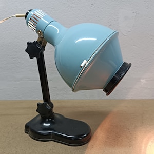 May include: A vintage blue and black desk lamp with a clamp base and a round shade. The lamp is turned on and the light is shining.