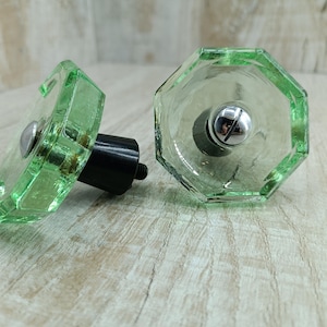Op de afbeelding: Twee groene glazen kastknoppen met zilveren metalen accenten. De knoppen hebben een facetgeslepen ontwerp en zijn bevestigd aan zwarte metalen bases.