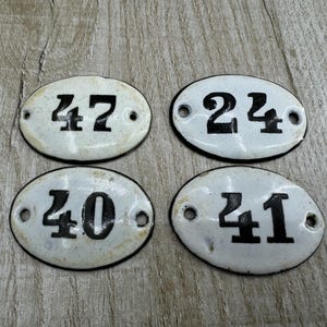 Könnte beinhalten: Vier weiße ovale Keramikfliesen mit schwarzen Zahlen. Die Zahlen sind 24, 40, 41 und 47.