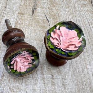 May include: Two wooden cabinet knobs with a floral design. The knobs have a dark brown finish with a clear, rounded top that encases a pink rose with green leaves. The knobs are set against a light wood surface.