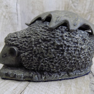 May include: A silver-coloured metal hedgehog figurine with a leaf-shaped lid. The hedgehog is lying down with its eyes closed and has a detailed textured surface.