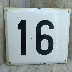 Könnte beinhalten: Weißes Email-Hausnummernschild mit schwarzen Zahlen, die Nummer 16.