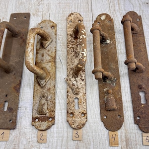 Peut inclure: Cinq poignées de porte antiques en métal rouillé de différents modèles. Chaque poignée a une plaque arrière rectangulaire avec un trou de serrure et des trous de montage. Les poignées sont numérotées de 1 à 5 sur de petites étiquettes en papier.