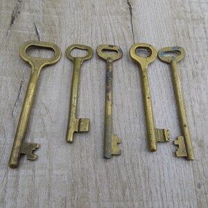 May include: Six antique brass keys with varying shapes and sizes. The keys are arranged horizontally on a light brown wooden surface.