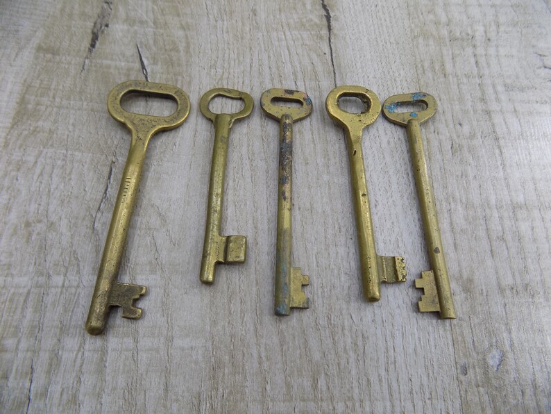 May include: Six antique brass keys with varying shapes and sizes. The keys are arranged horizontally on a light brown wooden surface.