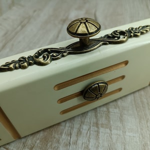 Op de afbeelding: Een witte lade met gouden rand en twee sierlijke messing knoppen. De knoppen hebben een decoratief bloemmotief.