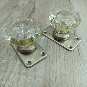 May include: Two antique glass door knobs with silver metal plates. The knobs are clear glass with a textured surface and have a raised design.