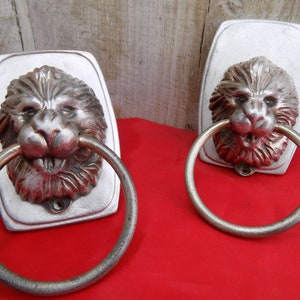 Op de afbeelding: Twee zilveren handdoekringen met een leeuwenkop op een witte achtergrond. De ringen zijn met schroeven aan de muur bevestigd.