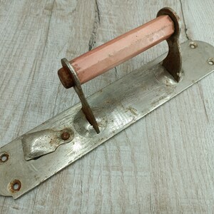 May include: A vintage metal door handle with a pink wooden knob. The handle is attached to a metal plate with two screws. The handle is rusty and shows signs of wear.