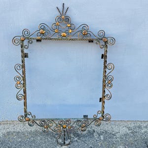 May include: Ornate, vintage-style metal frame with a square opening. The frame features intricate scrollwork, decorative accents, and a mix of gold and dark metal finishes. It is designed to hold a mirror or artwork.