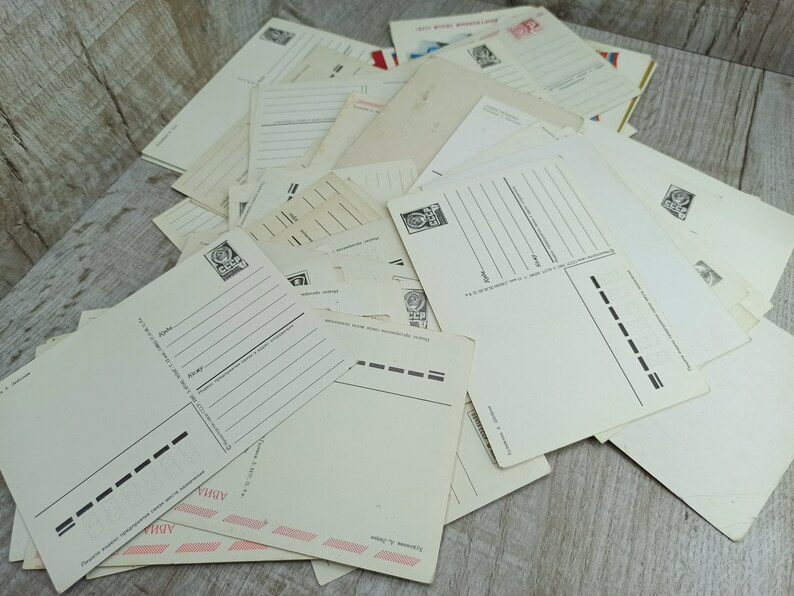 May include: A pile of vintage postcards, mostly cream-colored, with printed lines for writing. Some postcards have stamps and Cyrillic text. The postcards are arranged on a light-colored wooden surface, creating a nostalgic aesthetic.