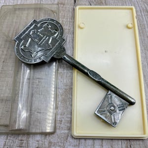 May include: A silver-coloured key with an elaborate design, placed on a cream plastic tray. The key features a detailed head and a square end. A clear plastic case is also visible.