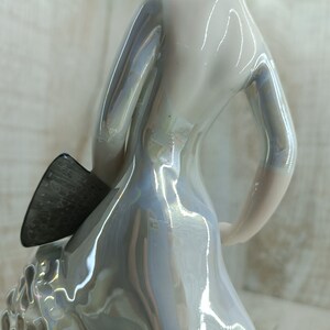 K&ouml;nnte beinhalten: Eine wei&szlig;e Keramikfigur einer Frau mit perlmuttartigem Finish. Die Frau tr&auml;gt einen flie&szlig;enden Rock und hat ihren Arm erhoben. Die Figur h&auml;lt ein kleines, dunkelgraues Objekt in ihrer Hand.