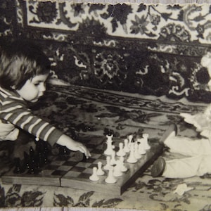 May include: A black and white photo of two children playing chess. The child on the left is wearing a striped shirt and the child on the right is wearing a long-sleeved shirt. The chessboard is in the center of the image.