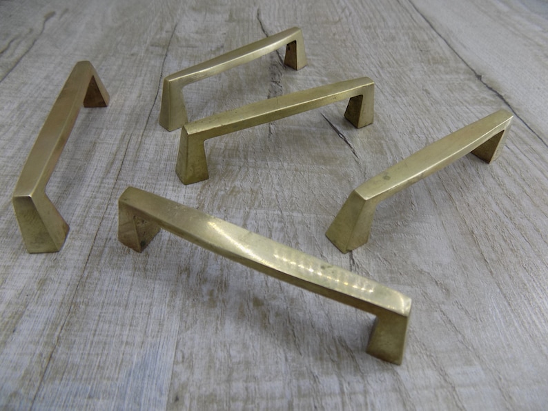 Op de afbeelding: Vijf messingkleurige metalen kastgrepen met een geometrisch ontwerp. De grepen zijn in een halve cirkel op een licht houten oppervlak gerangschikt.