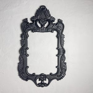 May include: Ornate black picture frame with a rectangular opening. The frame features elaborate scrollwork and floral details, with a decorative top and bottom. The frame is mounted on a textured white wall.