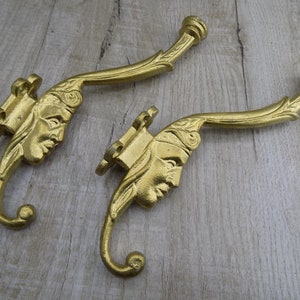 Puede incluir: Dos ganchos de metal de color dorado con diseños ornamentados que presentan una cara en cada gancho. Los ganchos son curvos y tienen un extremo redondeado.