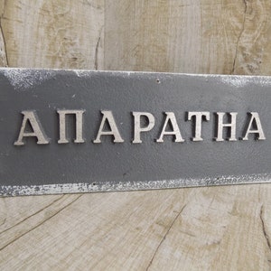 May include: A gray rectangular sign with white distressed paint around the edges. The word "APARATNA" is written in silver raised letters.